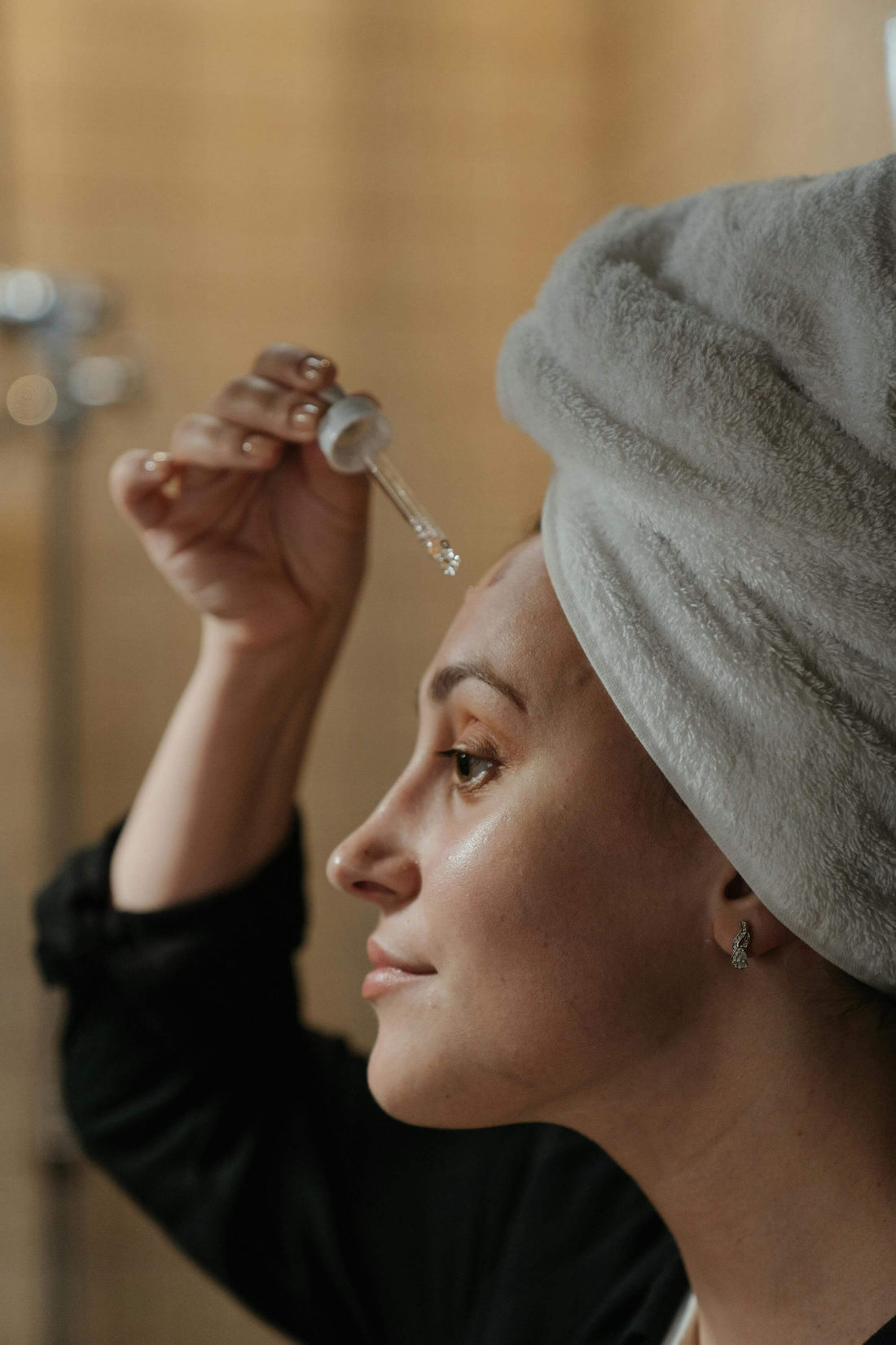 Woman applying hydrating face serum with a dropper, wrapped in a towel.