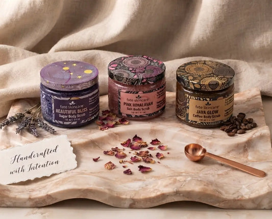 Three glass jars of Fate Skincare body scrubs—Beautiful Bliss Sugar Body Scrub, Pink Himalayan Salt Body Scrub, and Java Glow Coffee Scrub—displayed on a marble tray. 