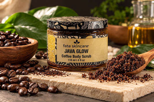 Java Glow Coffee Scrub™ by Fate Skincare in a 4 oz botanical-patterned jar, surrounded by roasted coffee beans, a wooden spoon of coffee scrub, and green leaves.